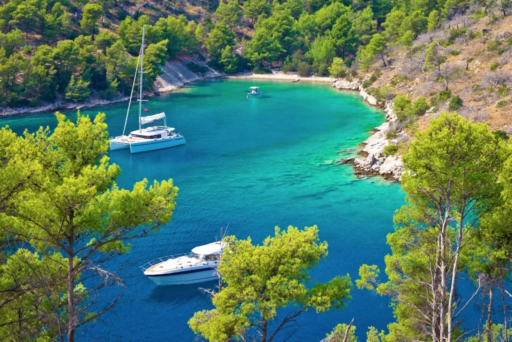 Bellissima baia di Brac in Croazia con acque cristalline e lussureggiante vegetazione verde. Due barche a vela, una più grande con un catamarano bianco e una più piccola, sono ancorate nel mare calmo.