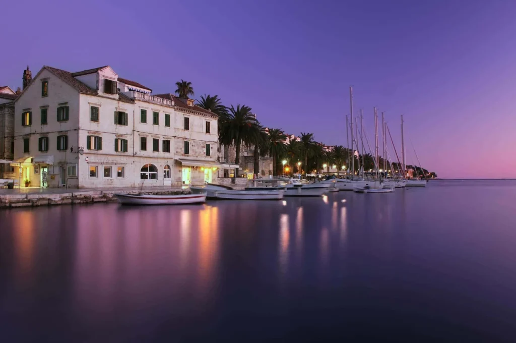 Tramonto mozzafiato sul lungomare della città di Hvar, Croazia, con una fila di barche a vela ormeggiate. L'acqua è calma e riflette le luci soffuse della sera, mentre le antiche facciate in pietra degli edifici si affacciano sul porto. Una immagine di serenità e di eleganza tipica della Croazia, anticipando ciò che rende queste crociere così incantevoli e ricercate.