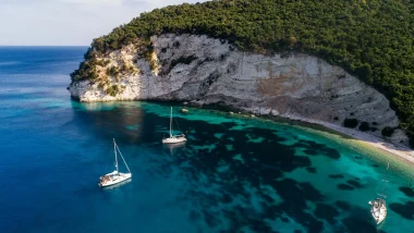 Barche a vela ancorate in una baia tranquilla di Atoko, vicino a Itaca, con acque trasparenti e scogliere coperte di vegetazione.