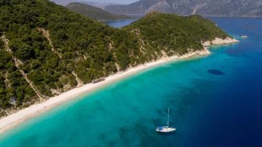 Barca a vela ancorata al largo di una spiaggia sabbiosa e incontaminata di Itaca, con colline verdi e acque turchesi.