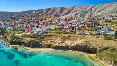 Vista aerea della spiaggia di Prika, pittoresca città costiera istriana con case colorate affacciate sulla baia tranquilla dalle acque di colore turchese intenso e cielo azzurro.