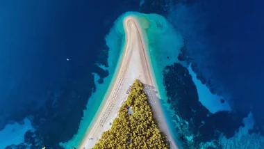 Vista aerea della Spiaggia di Bol Dalmazia, che mostra la caratteristica punta a forma di V fatta di ciottoli bianchi circondata dalle acque azzurre dell'Adriatico, con alberi di pino verdi rigogliosi ai lati della spiaggia e un parco acquatico galleggiante ancorato nelle vicinanze.