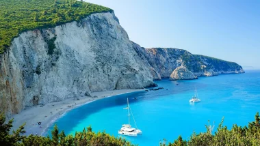 Catamarano bianco ancorato nelle acque turchesi presso la Spiaggia di Porto Katsiki, Lefkada, con imponenti scogliere calcaree e vegetazione lussureggiante. Vacanze in Barca a Vela alle Isole Ionie