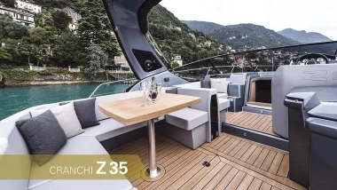 Salottino di Poppa della barca a motore Cranchi Z35 - Area lounge all'aperto con elegante tavolo da pranzo in legno, sullo sfondo dell'immagine montagne lussureggianti e le acque cristalline del mare.